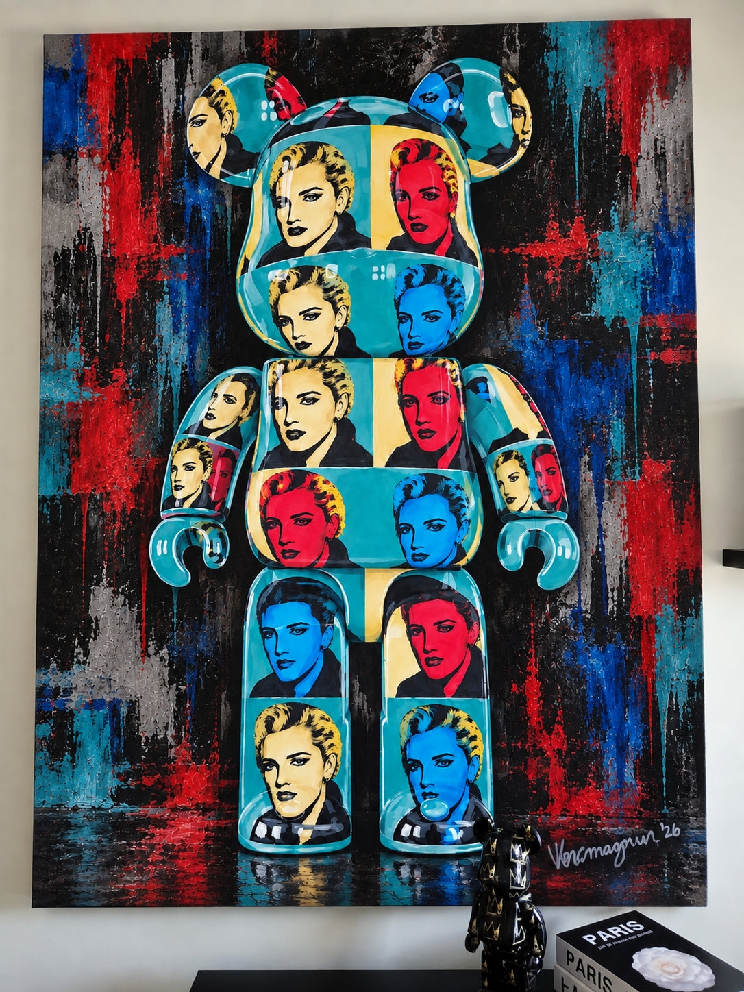 A large pop-art style bear-shaped canvas painting featuring multiple stylized portraits in blue, red, black, and cream tones, displayed against a white wall with a small figurine and books at the bottom.